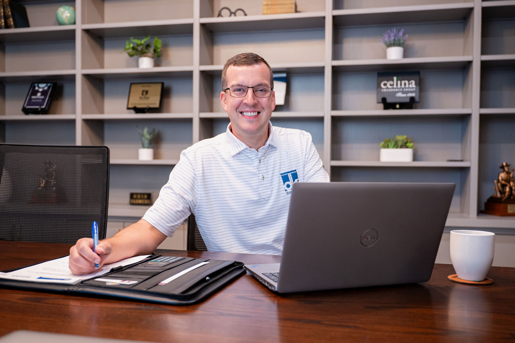 Smiling professional sitting at a desk with a laptop, writing notes in a modern office setting